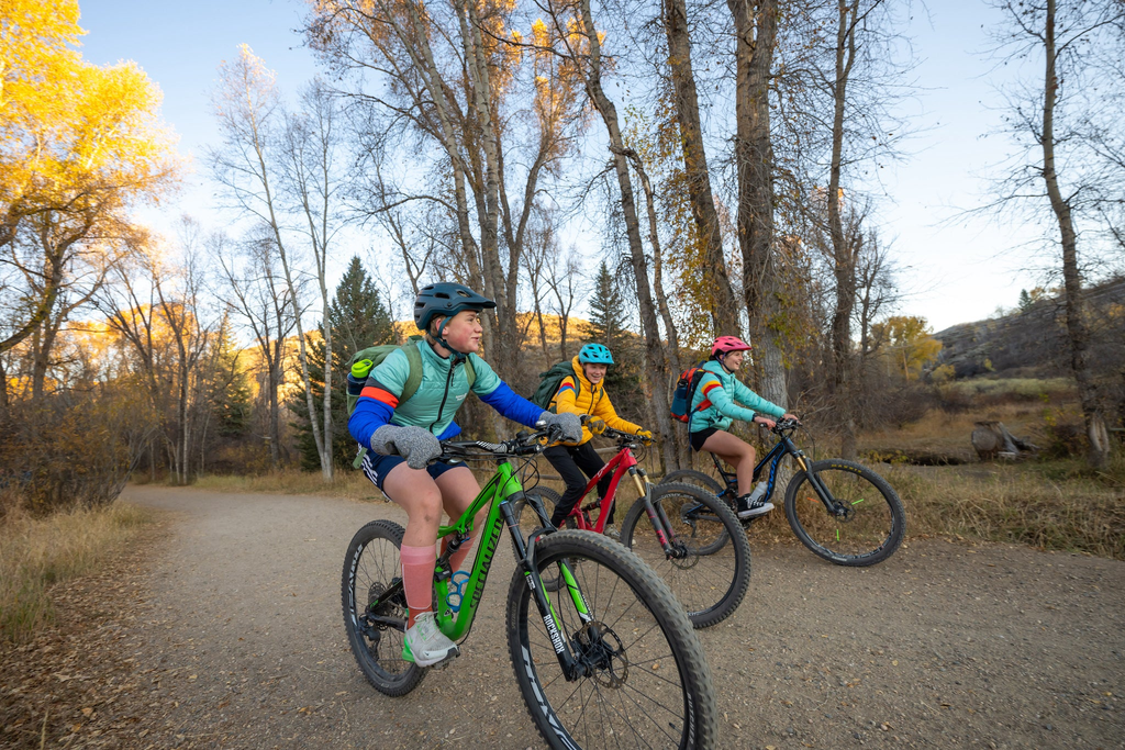 Three kids riding bikes to school on a dirt path with yellow fall leaves behind them, laughing, smiling and staying warm with Town Hall Outdoor Co jackets.