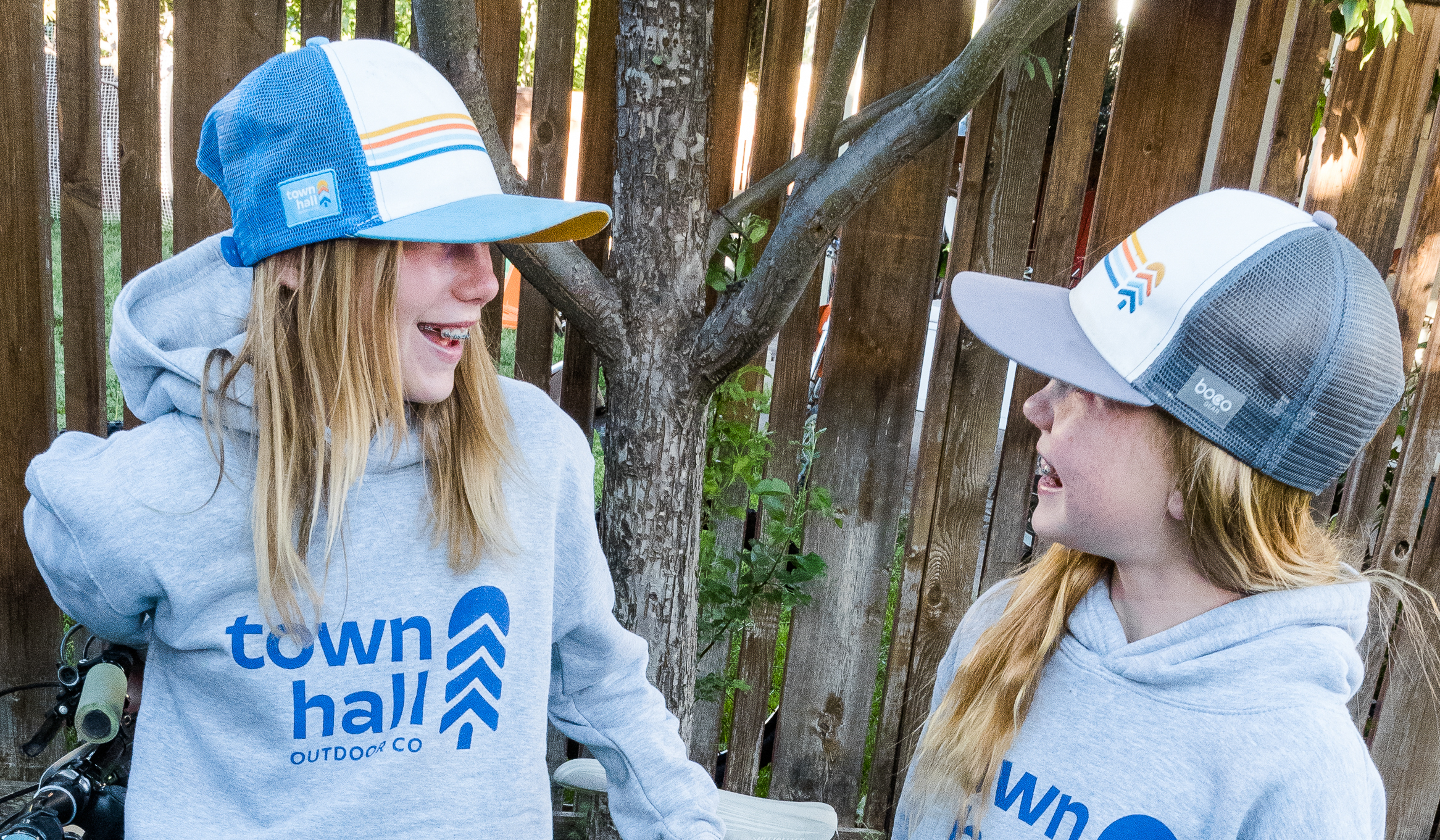 Two young girls are smiling and chatting by a fence on a backyard spring day, wearing their Town Hall kids hoodies and Trucker Hats.