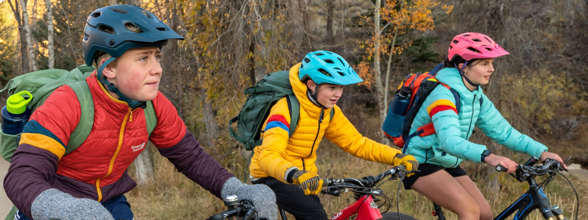 three kids riding bikes to school in colorful jackets with backpacks