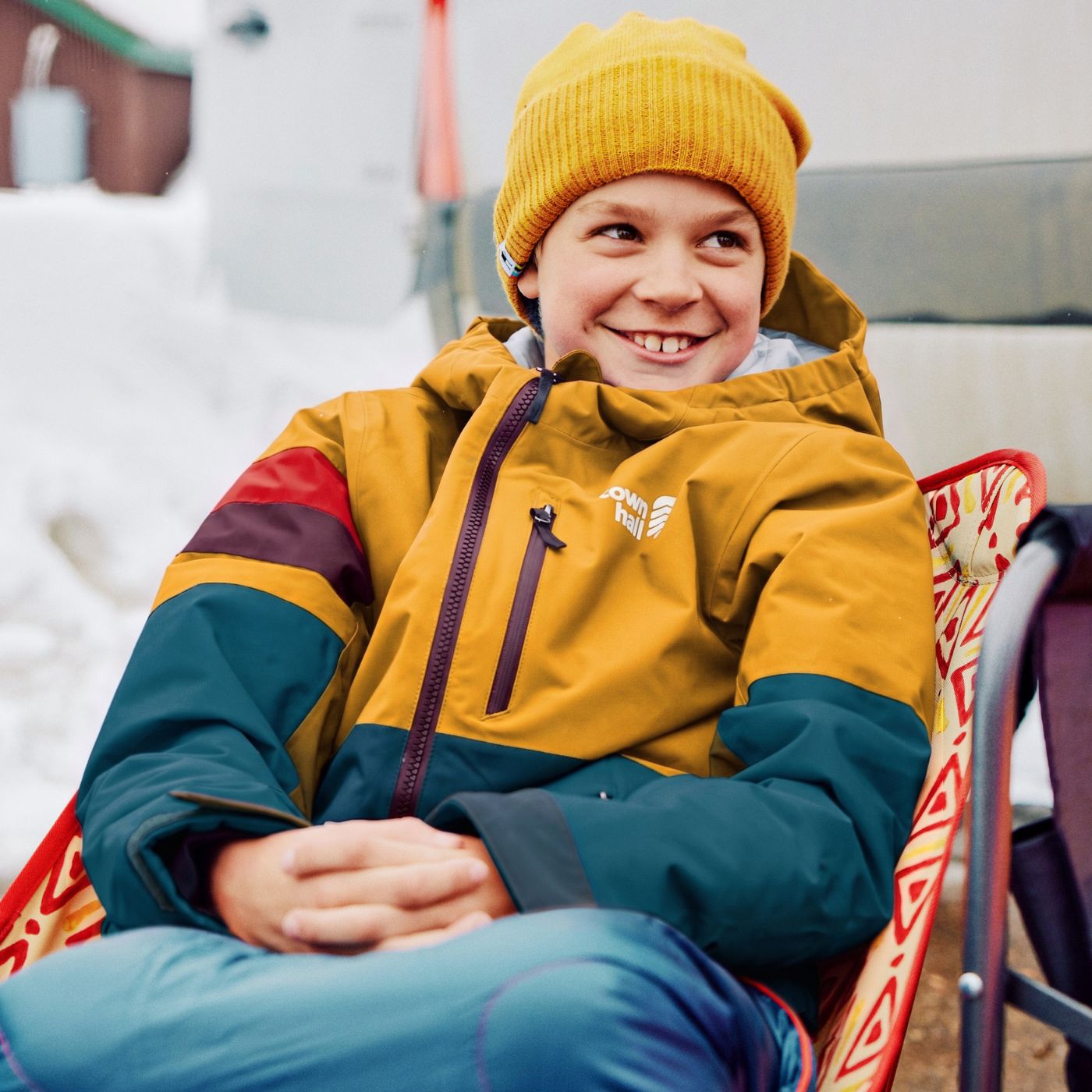 A smiling child wearing a mustard yellow knit beanie and a colorblock ski jacket in mustard yellow, teal, and red, sitting in a red patterned chair outdoors in a snowy setting.