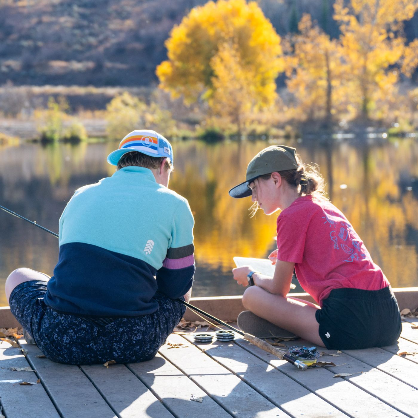 Two people sitting on a wooden dock by a lake, seen from behind, with one wearing a light blue and navy long-sleeve shirt and a blue cap, and the other wearing a pink t-shirt and a green cap. Trees with yellow autumn foliage are reflected in the water in the background.