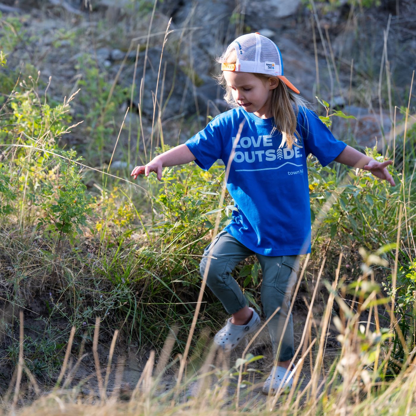 A young girl wearing a blue t-shirt with 'LOVE OUTSIDE' printed on the front, olive green pants, and a white trucker hat with orange trim, walking through tall grass outdoors.