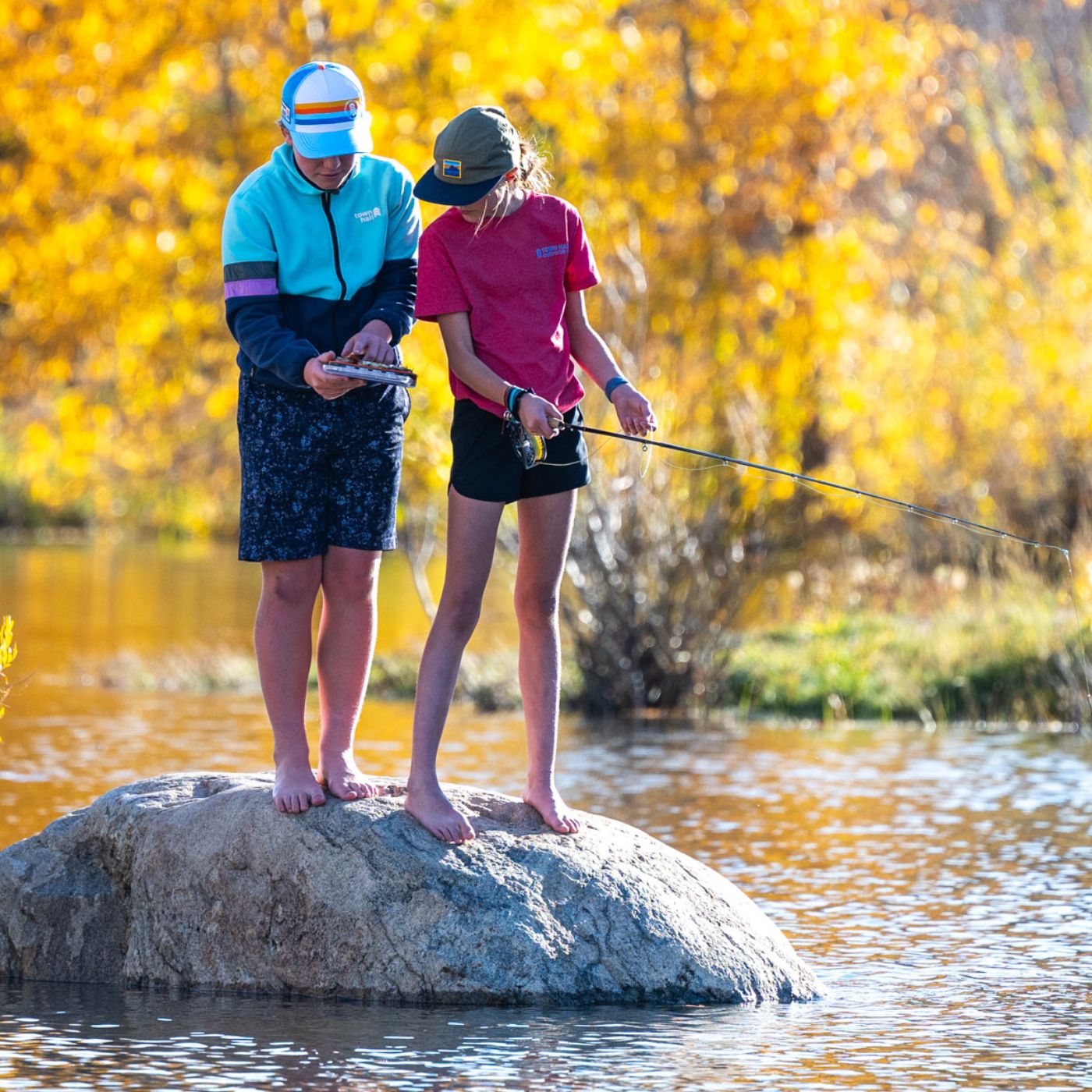 Two people fishing on a rock in a river with autumn trees in the background