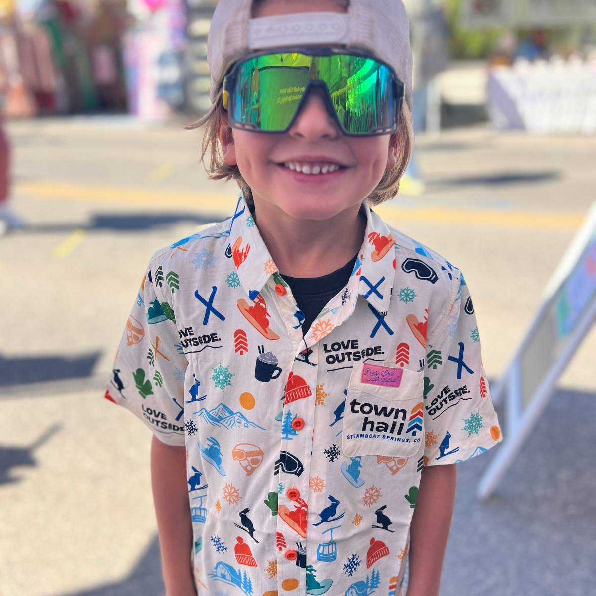 Child wearing a colorful shirt with graphics and reflective sunglasses in an outdoor setting.