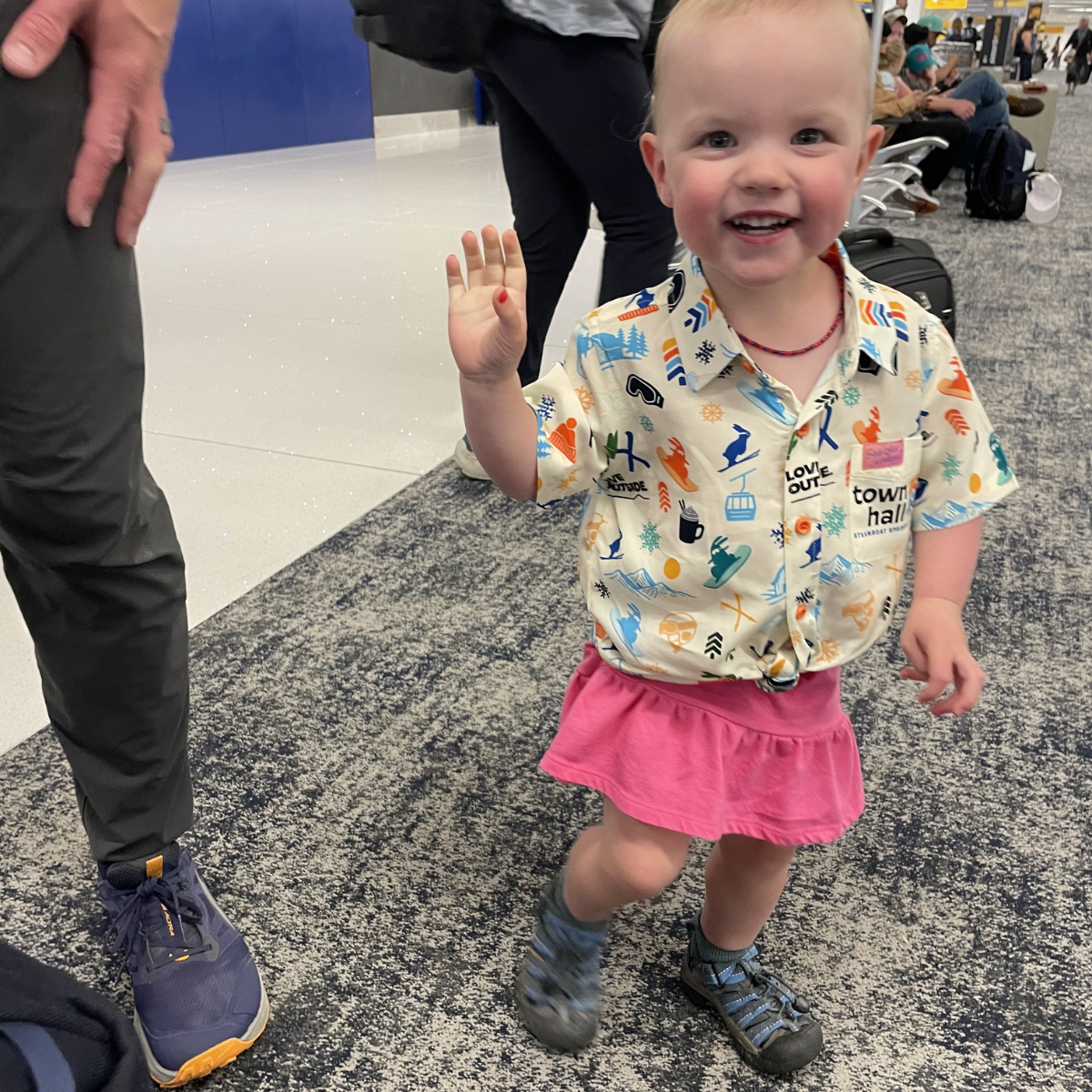 Child in a colorful shirt and pink skirt waving at the camera in an indoor setting.