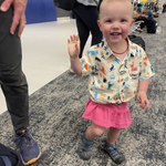 Child in a colorful shirt and pink skirt waving at the camera in an indoor setting.