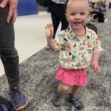 Child in a colorful shirt and pink skirt waving at the camera in an indoor setting.