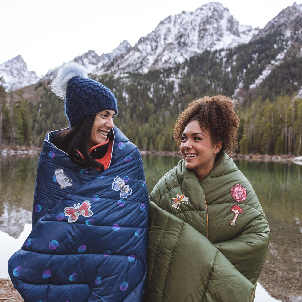 Two outdoorsy women are sitting under down blankets covered in NOSO patches with snow covered mountains behind them