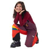 Child kneeling in a Grape Jam purple and orange Mountain Town kids snow bib with black ankle panels and ski boots.