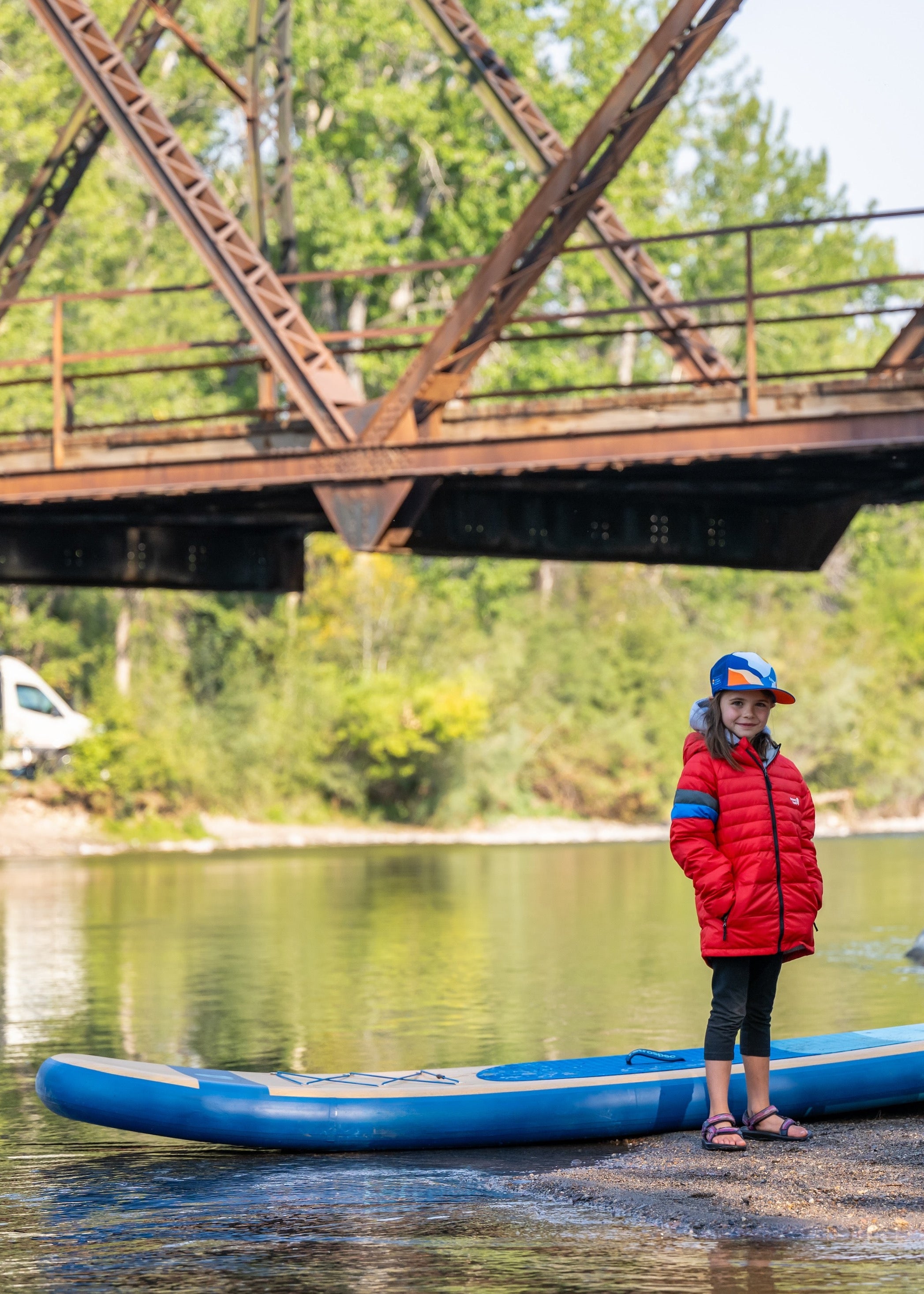 A person in a red puffer jacket and blue cap stands on a riverbank under a large rusty metal bridge, holding a blue canoe. Rocks, green trees, and calm water are visible in the background.