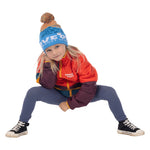 Child crouching in the blue Kids Love Outside Beanie with striped crown and pom-pom, wearing a colorblock puffy jacket.