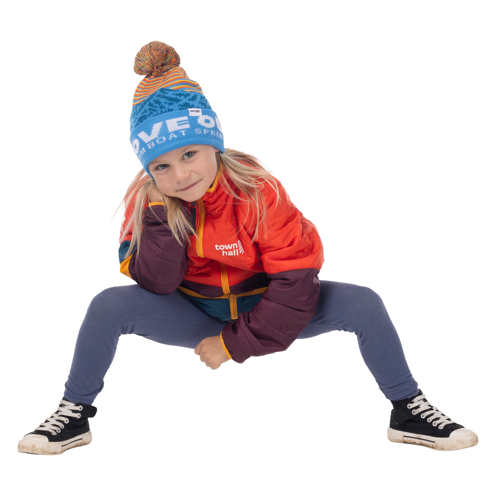 Child crouching in the blue Kids Love Outside Beanie with striped crown and pom-pom, wearing a colorblock puffy jacket.