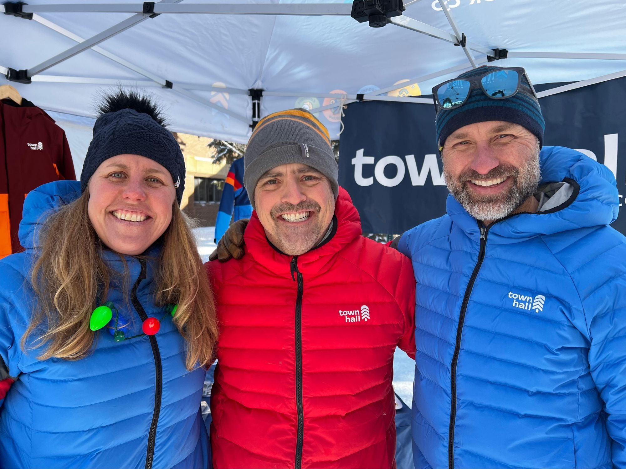 Three people wearing blue and red jackets with a visible 'town hall' logo, standing under a white tent.