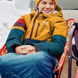 A smiling child wearing a mustard yellow and teal colorblock ski jacket, a yellow beanie, and blue pants, sitting on a red folding chair outdoors.