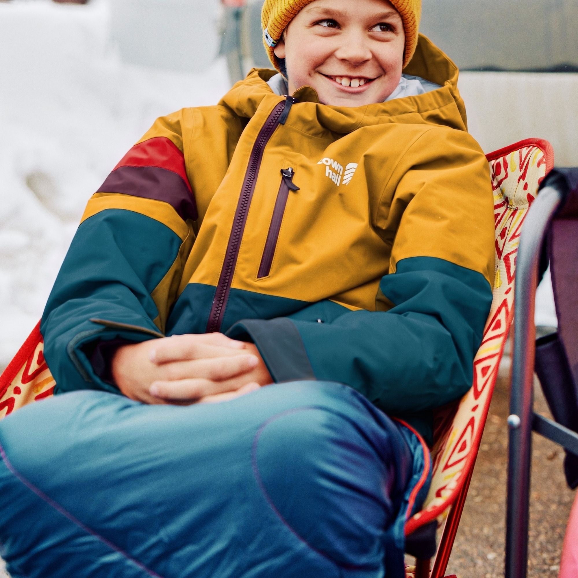 A smiling child wearing a mustard yellow and teal colorblock ski jacket, a yellow beanie, and blue pants, sitting on a red folding chair outdoors.