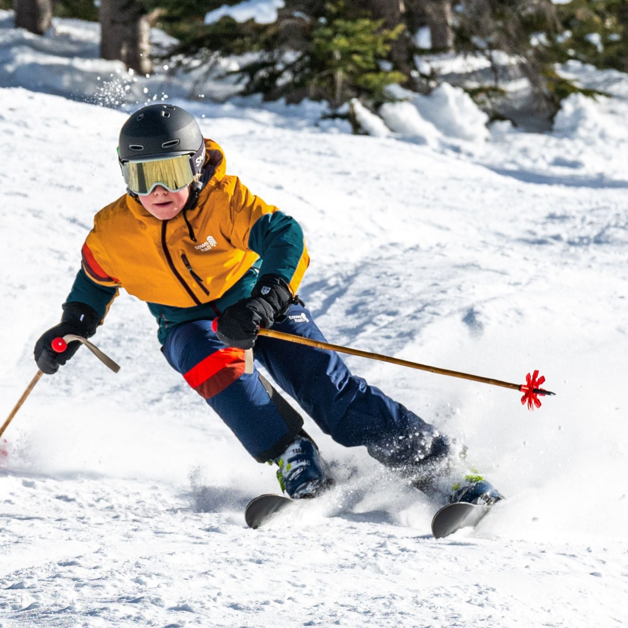 A child skier in a bright yellow and teal jacket, blue pants, and a black helmet with goggles carving through fresh powder on a snowy mountain.