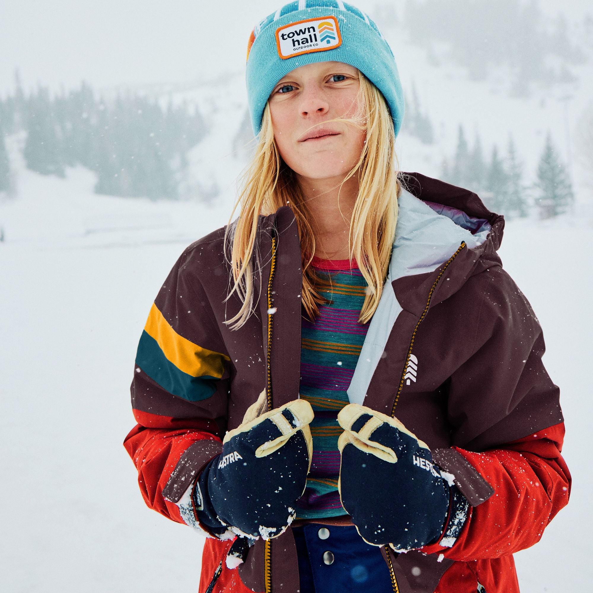 A young girl wearing a turquoise Town Hall beanie, a dark brown and red ski jacket, and dark blue gloves, standing in a snowy outdoor setting.