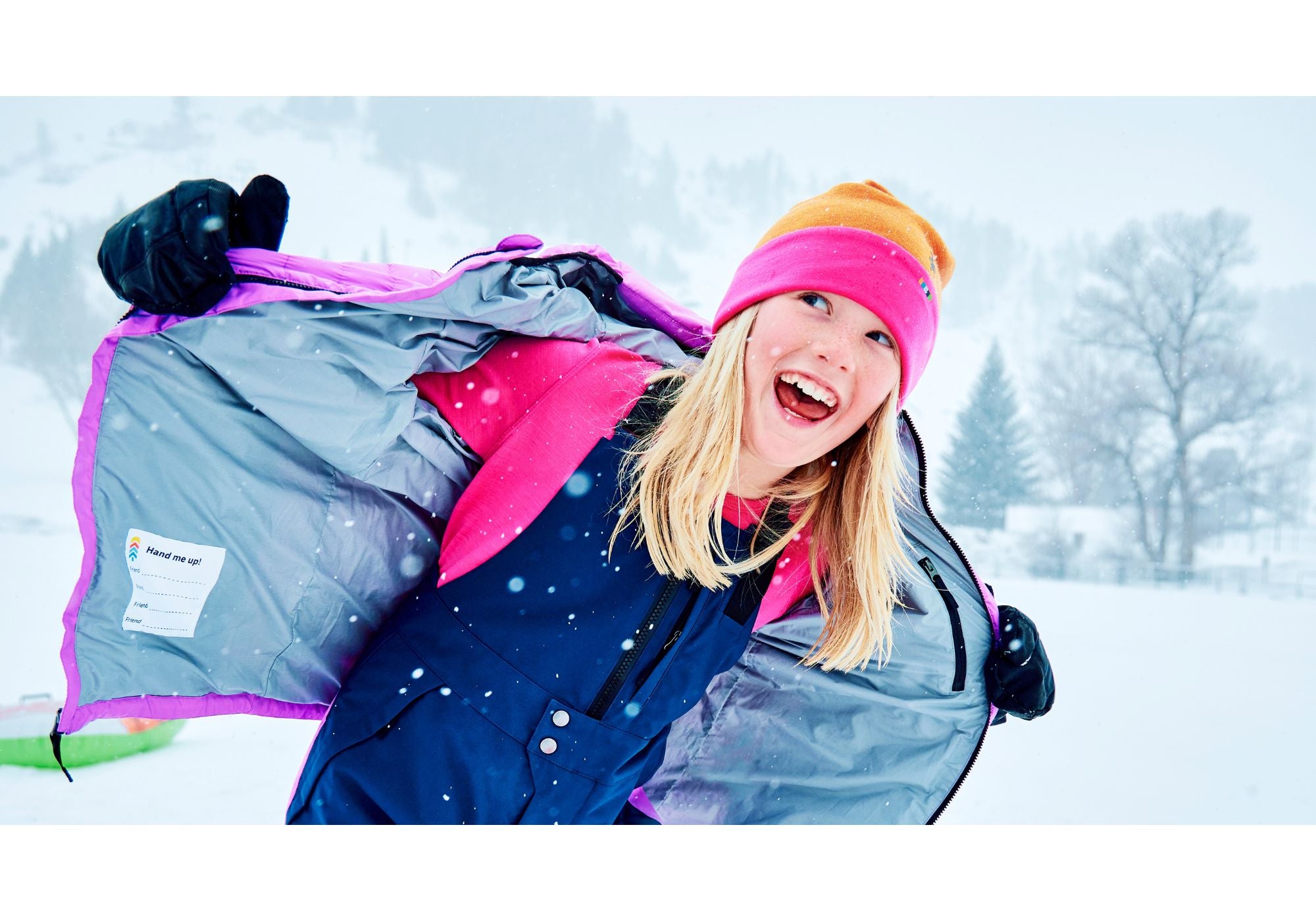 A girl in a snowy outdoor setting wearing a navy blue ski jacket with a bright pink and purple lining, a pink and orange beanie, and black gloves, smiling as she spreads the jacket open.